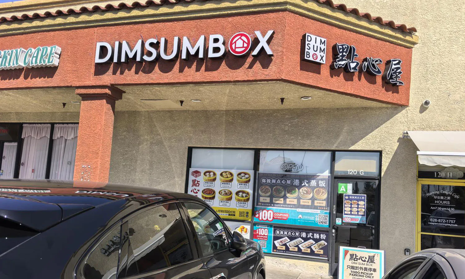Corner storefront of DIMSUMBOX dim sum restaurant in sunlight, orange sign, dim sum posters on windows, black SUV parked in front, strip mall scene.
